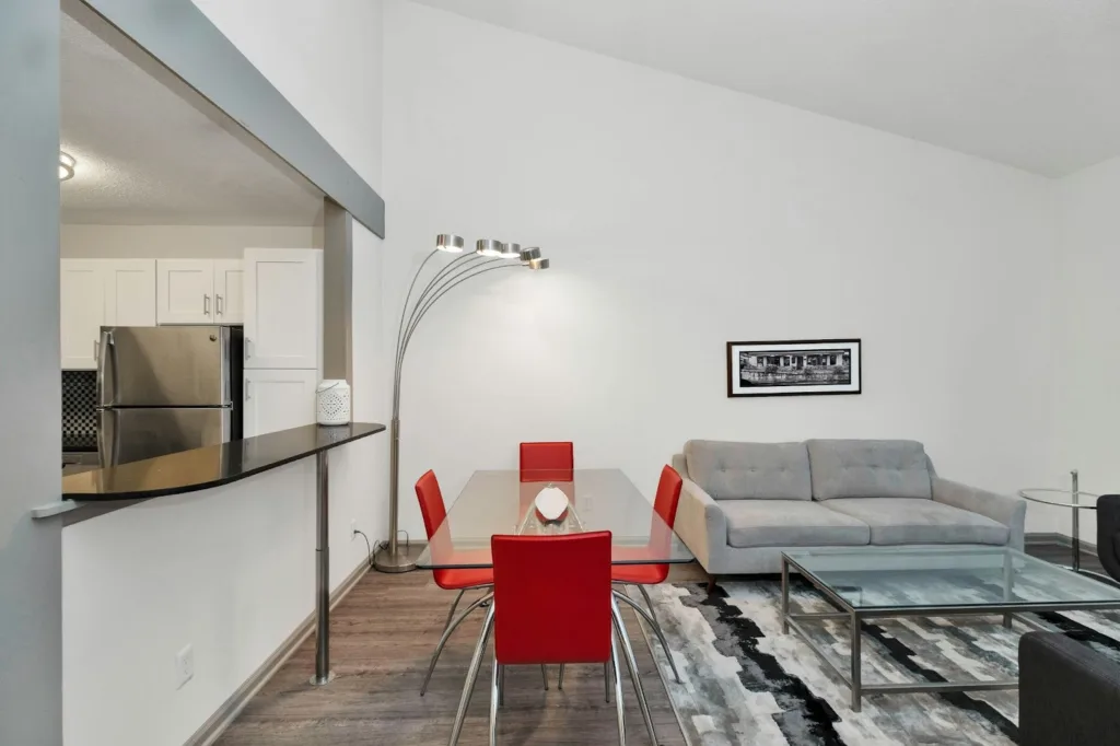 Neutral open-plan living room with a red dining set beside a grey sofa and patterned rug.