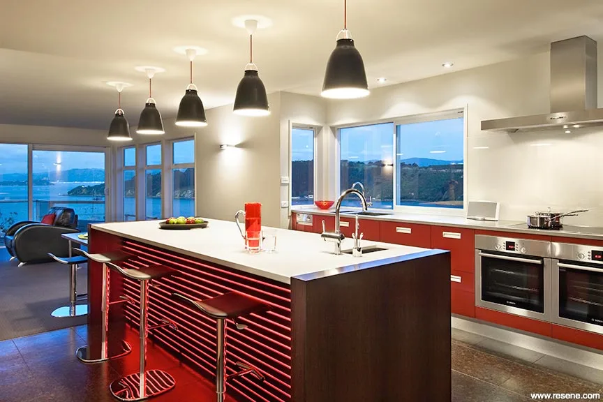 Modern kitchen with a white island and red cabinetry, pendant lights, and large windows at dusk