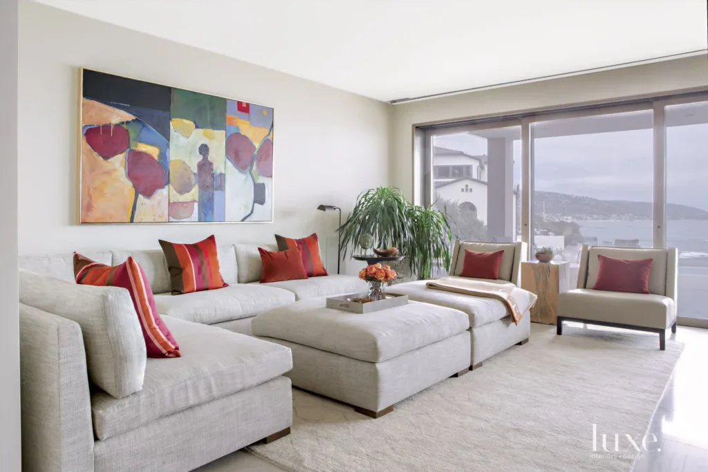 Neutral living room with a light grey sectional and red accent pillows, artwork on the wall, and a large window view.